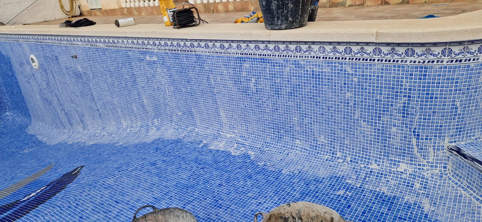 Vaso vacío de una piscina de gresite azul en obra, con restos de lechada en las paredes y herramientas en el borde